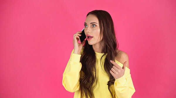 astonished young adult woman with open mouth speaking on cellphone isolated on pink  