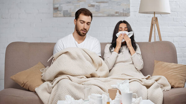 sick woman sneezing in tissue near man and bottles with pills on coffee table 