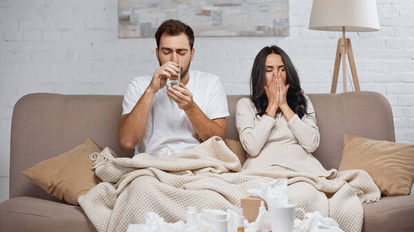 man drinking water near sick woman sneezing in living room 