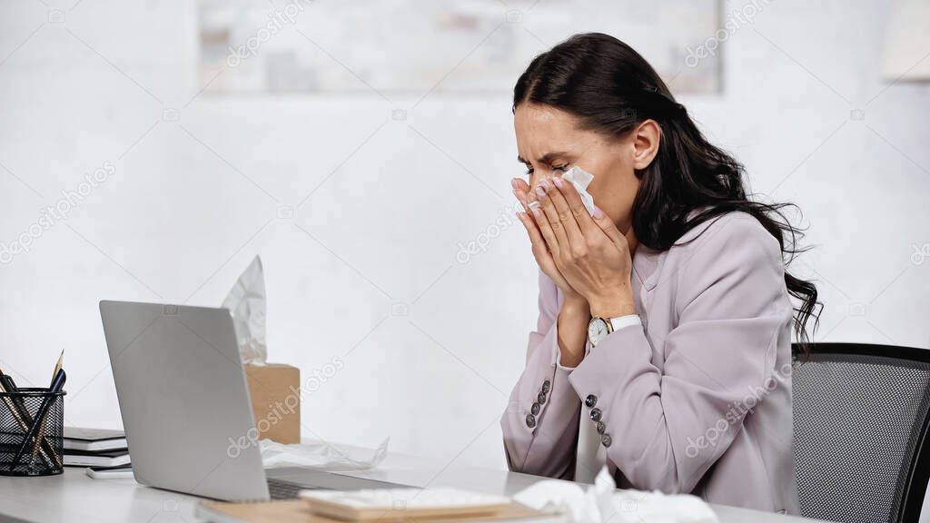 allergic woman sneezing in tissue near laptop on desk in office