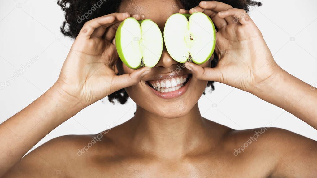 Smiling african american woman with naked shoulders holding pieces of apple near eyes isolated on grey 