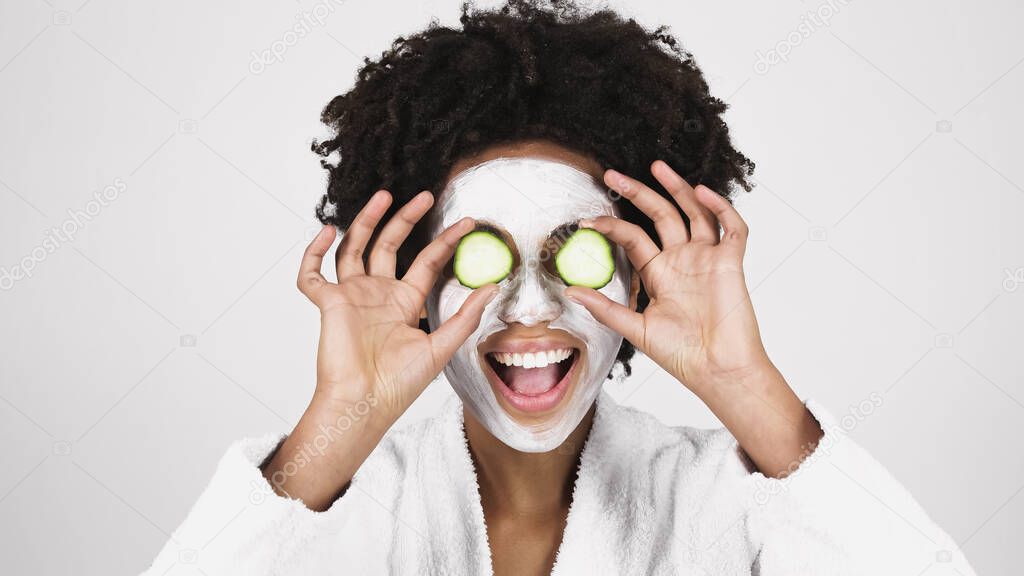 Excited african american woman in face mask holding cucumber slices near eyes isolated on grey 