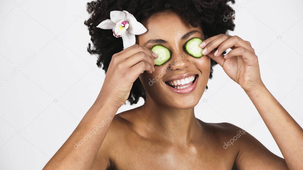 Cheerful african american woman with orchid flower in hair holding cucumber slices near eyes isolated on grey 