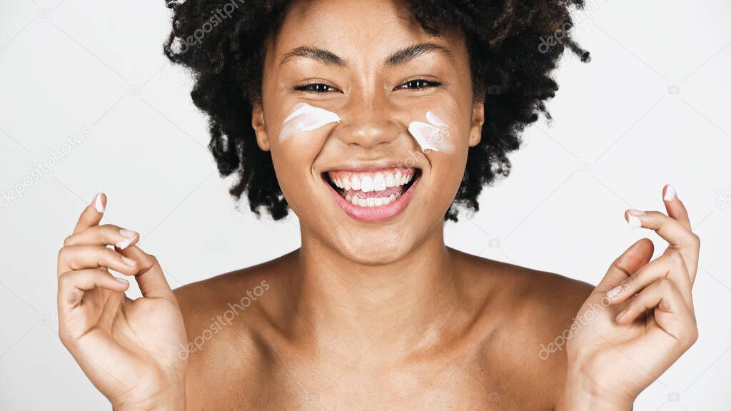 Positive african american woman with cream on face and naked shoulders looking at camera isolated on grey 