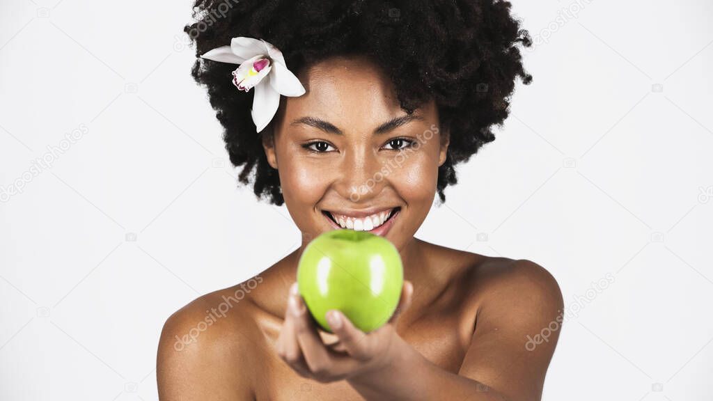Positive african american woman with orchid in hair holding apple on blurred foreground isolated on grey 
