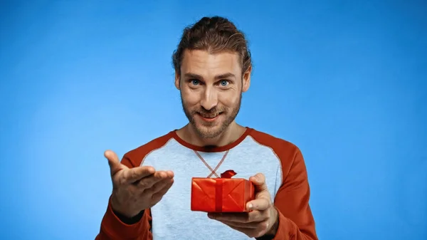 Hombre barbudo feliz sosteniendo regalo envuelto y señalando con la mano en azul - foto de stock