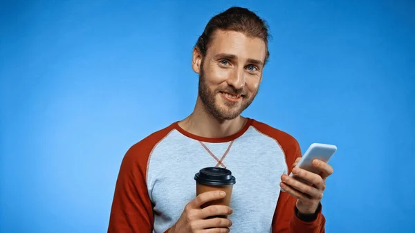 Веселий бородатий чоловік використовує смартфон, тримаючи паперову чашку на синьому — стокове фото