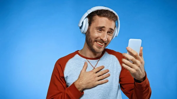 Homme heureux dans les écouteurs sans fil regardant le téléphone sur bleu — Photo de stock