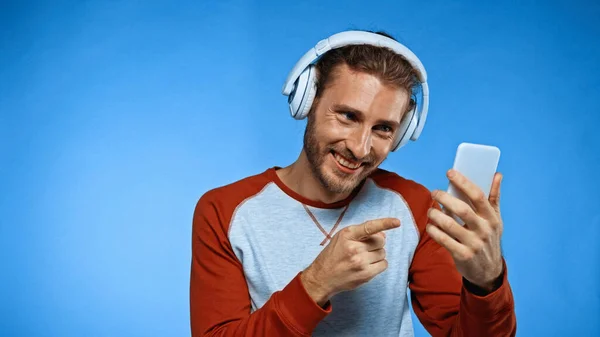 Homme heureux dans les écouteurs sans fil pointant avec le doigt au téléphone sur bleu — Photo de stock