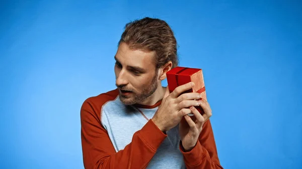 Neugieriger junger Mann schüttelt verpacktes rotes Geschenk auf blauem Grund — Stockfoto