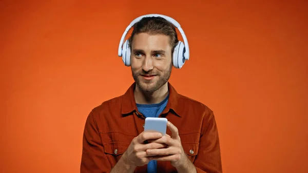 Радісний молодий чоловік в бездротових навушниках, використовуючи смартфон на помаранчевому — стокове фото