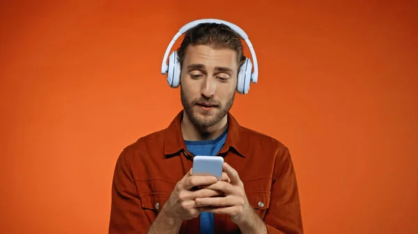 Bearded young man in wireless headphones using smartphone on orange — Stock Photo