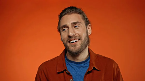 Hombre contento y barbudo sonriendo mientras mira hacia otro lado en naranja - foto de stock