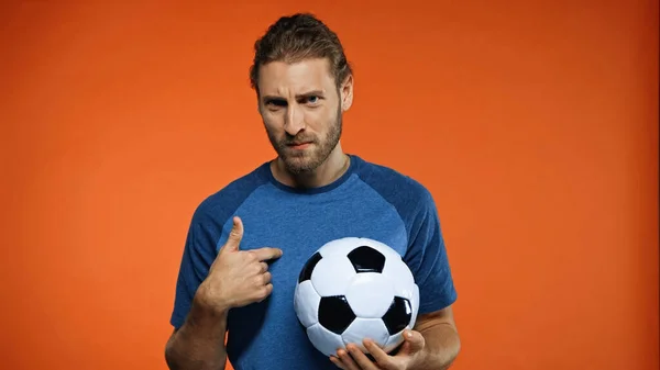 Football fan in blue t-shirt holding soccer ball and pointing at himself on orange — Stock Photo