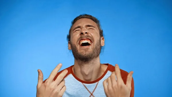 Молодий бородатий чоловік із закритими очима жестикулює під час сміху на синьому — стокове фото