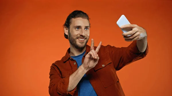 Barbudo joven mostrando signo de paz mientras toma selfie en naranja - foto de stock