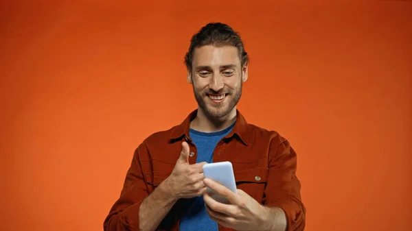 Bärtiger junger Mann zeigt Daumen nach oben, während er Selfie auf orange macht — Stockfoto