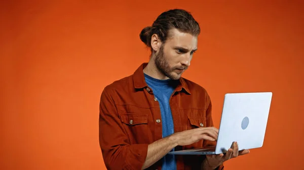 Bearded young man using laptop on orange — Stock Photo