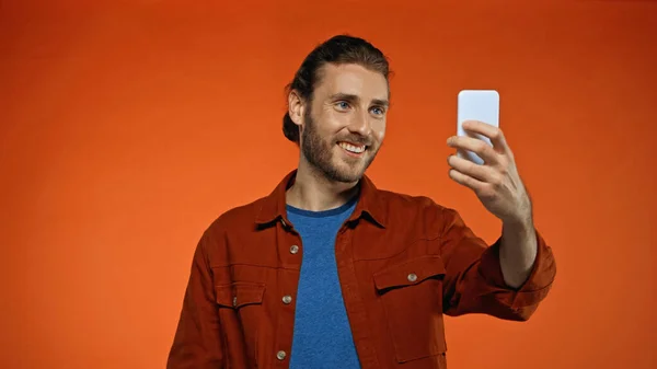 Hombre alegre sonriendo mientras mira el teléfono inteligente y tomar selfie en naranja - foto de stock
