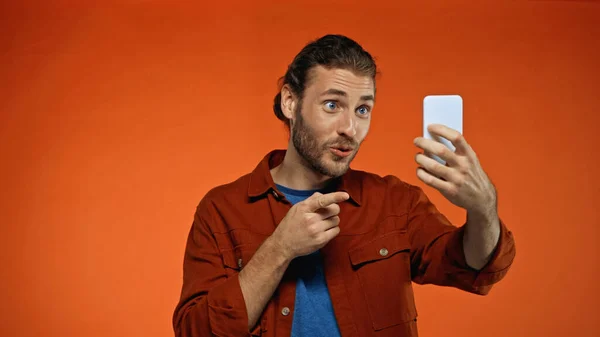 Homem alegre apontando com o dedo enquanto olha para o smartphone e tomar selfie em laranja — Fotografia de Stock