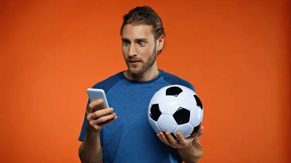 Tifoso sorpreso di calcio in t-shirt blu utilizzando smartphone e tenendo palla da calcio su arancione — Foto stock