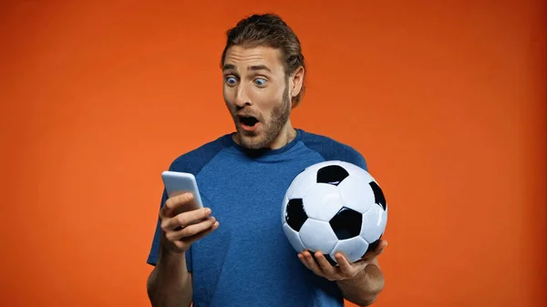 Шокований футбольний вентилятор у синій футболці за допомогою смартфона і тримає футбольний м'яч на помаранчевому — стокове фото