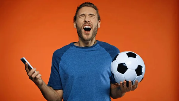 Abanico de fútbol en camiseta azul sosteniendo teléfono inteligente y pelota de fútbol mientras grita en naranja - foto de stock