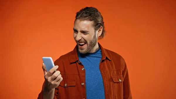 Homem barbudo animado usando telefone celular no fundo laranja — Fotografia de Stock