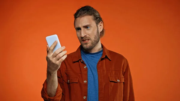 Dissatisfied young man looking at mobile phone on orange — Stock Photo