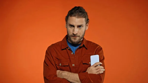 Bearded man standing with crossed arms and holding phone on orange — Stock Photo