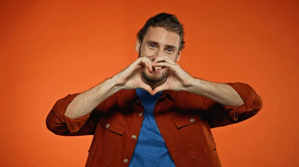 Joven barbudo sonriendo y mostrando el signo del corazón en naranja - foto de stock