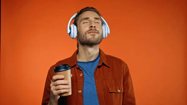 Pleased man in wireless headphones holding paper cup and listening music on orange — Stock Photo