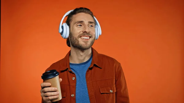 Cheerful man in wireless headphones holding paper cup and listening music on orange — Stock Photo