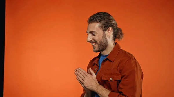 Positive young and bearded man smiling and standing with praying hands on orange — Stock Photo