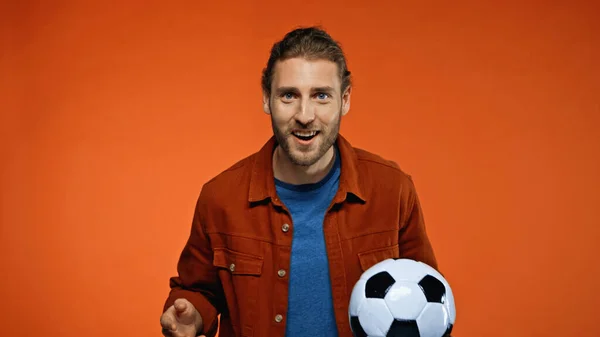 Позитивный футбольный фанат смотрит в камеру, держа футбольный мяч на оранжевом — стоковое фото