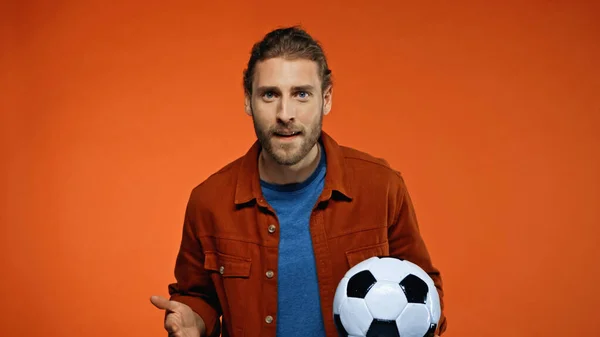 Football fan looking at camera while holding soccer ball and cheering on orange — Stock Photo