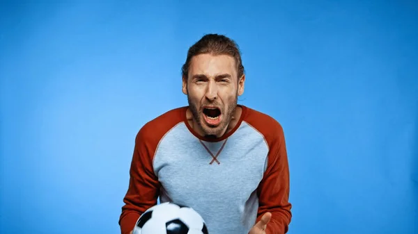 Расстроен молодой футбольный фанат держит футбольный мяч и кричит на синий — стоковое фото