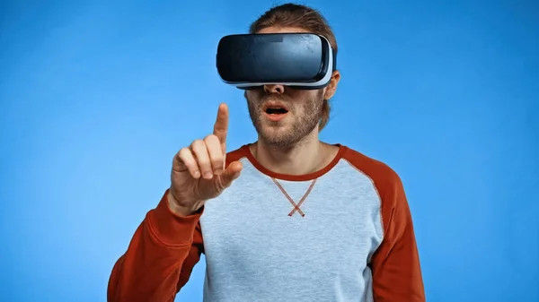 Bearded young man in virtual reality headset pointing with finger on blue — Stock Photo