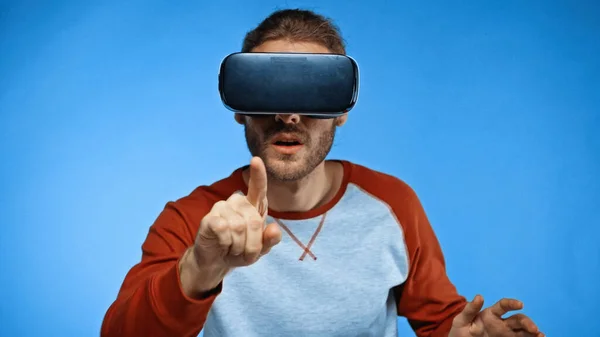 Bearded young man in virtual reality headset pointing with finger on blue — Stock Photo