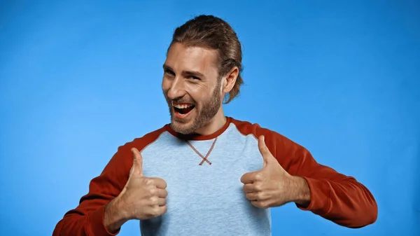 Positive young man showing thumbs up while looking at camera on blue — Stock Photo