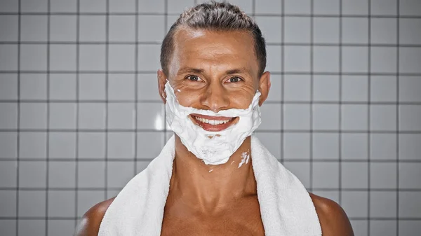 Sonriente hombre con espuma de afeitar en la cara mirando a la cámara en el baño - foto de stock