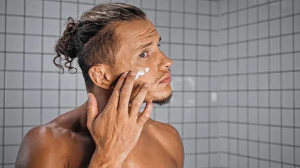 Без сорочки чоловік наносить крем для обличчя у ванній — стокове фото