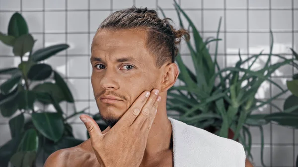 Shirtless man touching face near green plants on blurred background in bathroom — Stock Photo