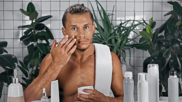 Homem sem camisa aplicando creme facial perto de plantas verdes no fundo borrado no banheiro — Fotografia de Stock