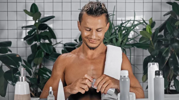 Happy man holding bottle with shaving foam near green plants on blurred background in bathroom — Stock Photo