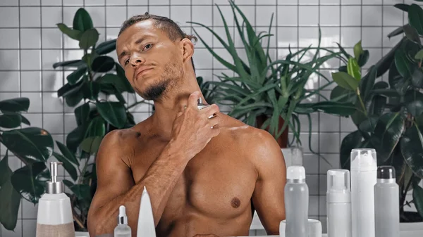 Hombre sin camisa aplicando perfume cerca de plantas verdes sobre fondo borroso en el baño - foto de stock