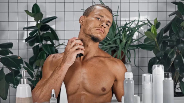 Uomo senza maglietta spruzzando profumo vicino piante verdi su sfondo sfocato in bagno — Foto stock