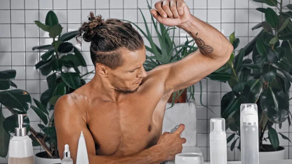 Tattooed man spraying deodorant near green plants on blurred background in bathroom — Stock Photo