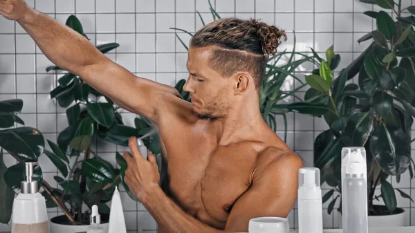 Uomo senza maglietta spruzzando deodorante vicino a piante verdi su sfondo sfocato in bagno — Foto stock