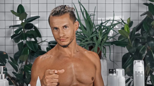 Uomo senza maglietta che punta con il dito mentre guarda la fotocamera vicino a piante verdi su sfondo sfocato in bagno — Foto stock